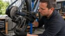 A cyclist inspects a bicycle chain and rear cassette in a workshop.