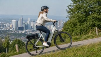 A helmeted rider climbing a paved hill on a city e-bike above an urban skyline.