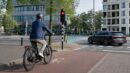 A helmeted city e-bike rider approaches an intersection while a car passes at a safe distance.