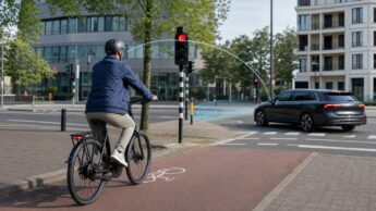 A helmeted city e-bike rider approaches an intersection while a car passes at a safe distance.