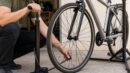 A rider uses a floor pump on a hybrid bike tire in a driveway workshop setting.