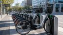 A row of unbranded docked city bike-share e-bikes charging at an urban transit stop.